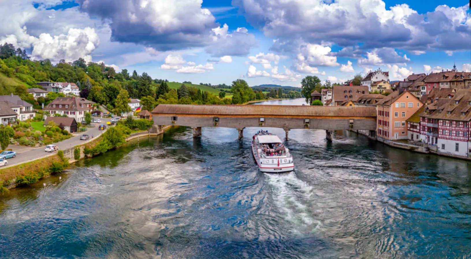 A sightseeing boat sails beneath a historic wooden bridge on the Rhine River, surrounded by colorful traditional Swiss houses and rolling green hills under a blue sky. This picturesque moment showcases the serene side of Swiss travel, making it an ideal highlight in the Swiss Travel Pass Guide for exploring Switzerland by boat and rail.