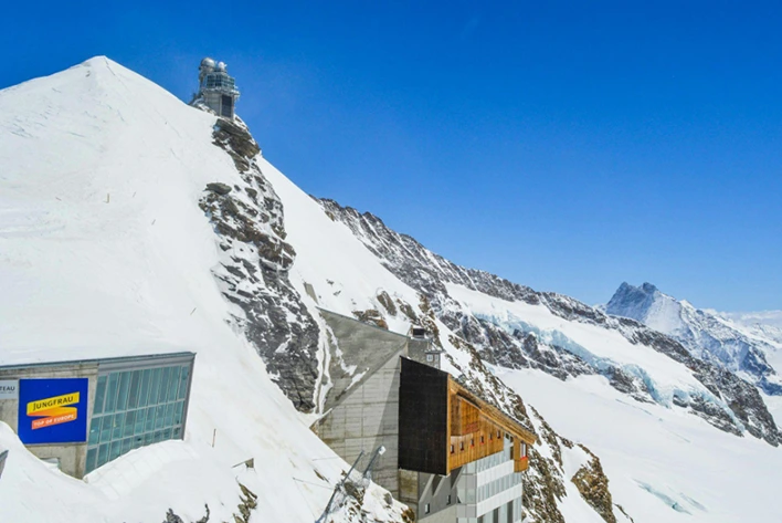 Scenic view of the Sphinx Observatory set against snowy mountains under a clear blue sky, with an old building in the foreground, capturing the stunning alpine landscape of Jungfraujoch, Switzerland.